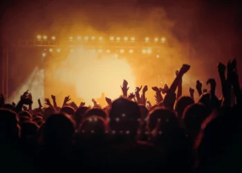 Koncert muzyczny. Pomarańczowe światła i dym unoszący się ze sceny. Ludzie unoszą ręce do góry.