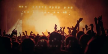 Koncert muzyczny. Pomarańczowe światła i dym unoszący się ze sceny. Ludzie unoszą ręce do góry.